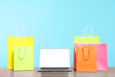 Photo of Internet shopping. Laptop and colorful paper bags on wooden table against light blue background