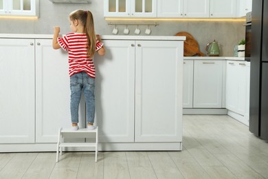 Photo of Little girl standing on step stool and reaching towards counter in kitchen, back view. Space for text