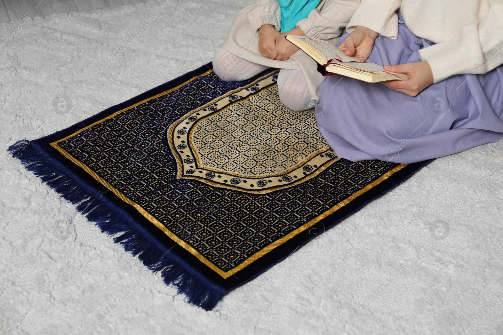 Photo of Muslim woman and her daughter with Quran praying on mat at home, closeup