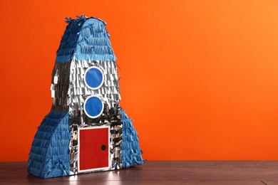 Photo of Bright pinata in shape of spaceship on table against orange background. Space for text