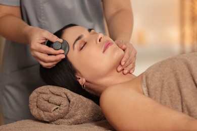 Photo of Young woman receiving facial massage with spa stone in salon