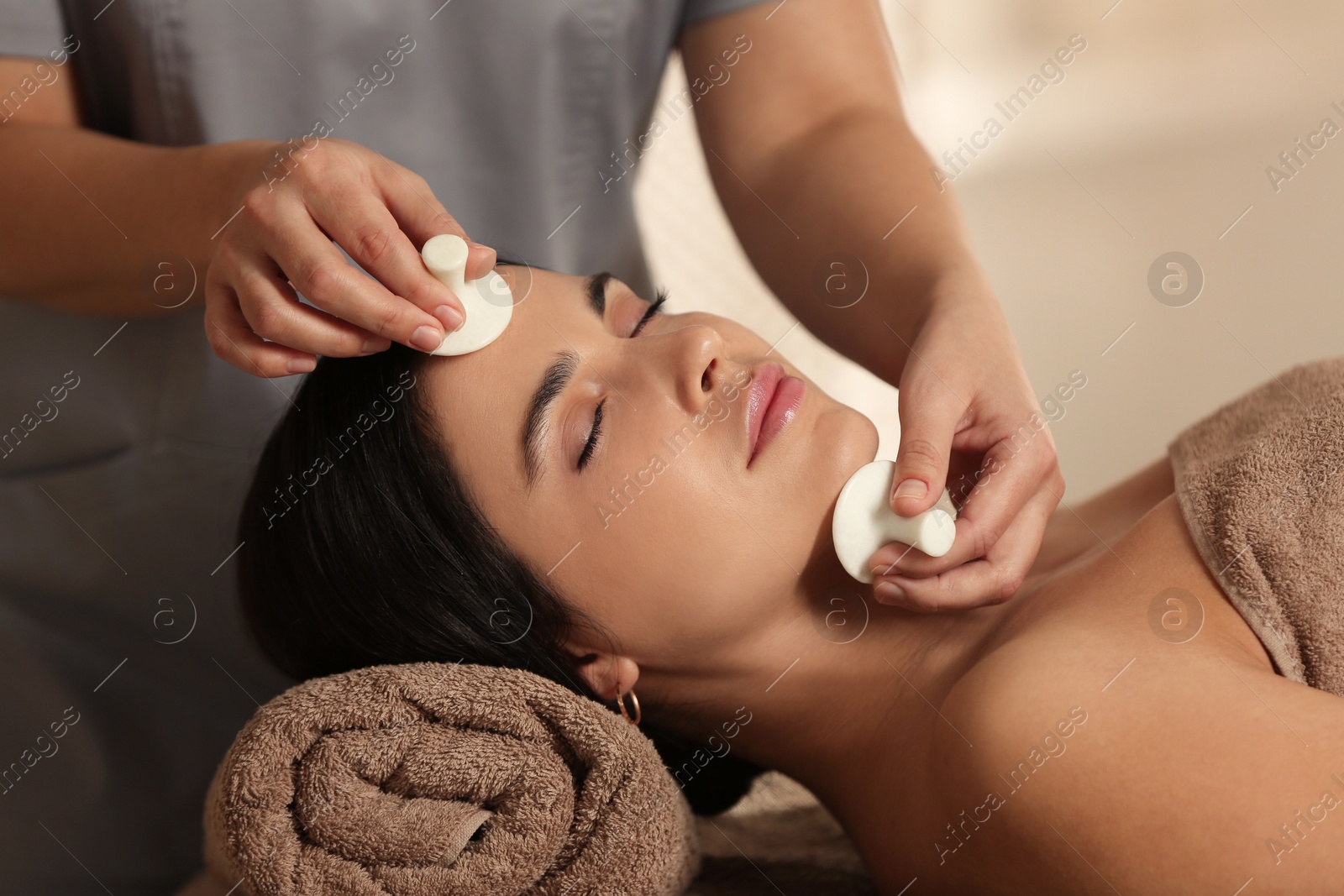 Photo of Young woman receiving facial massage with spa stones in salon