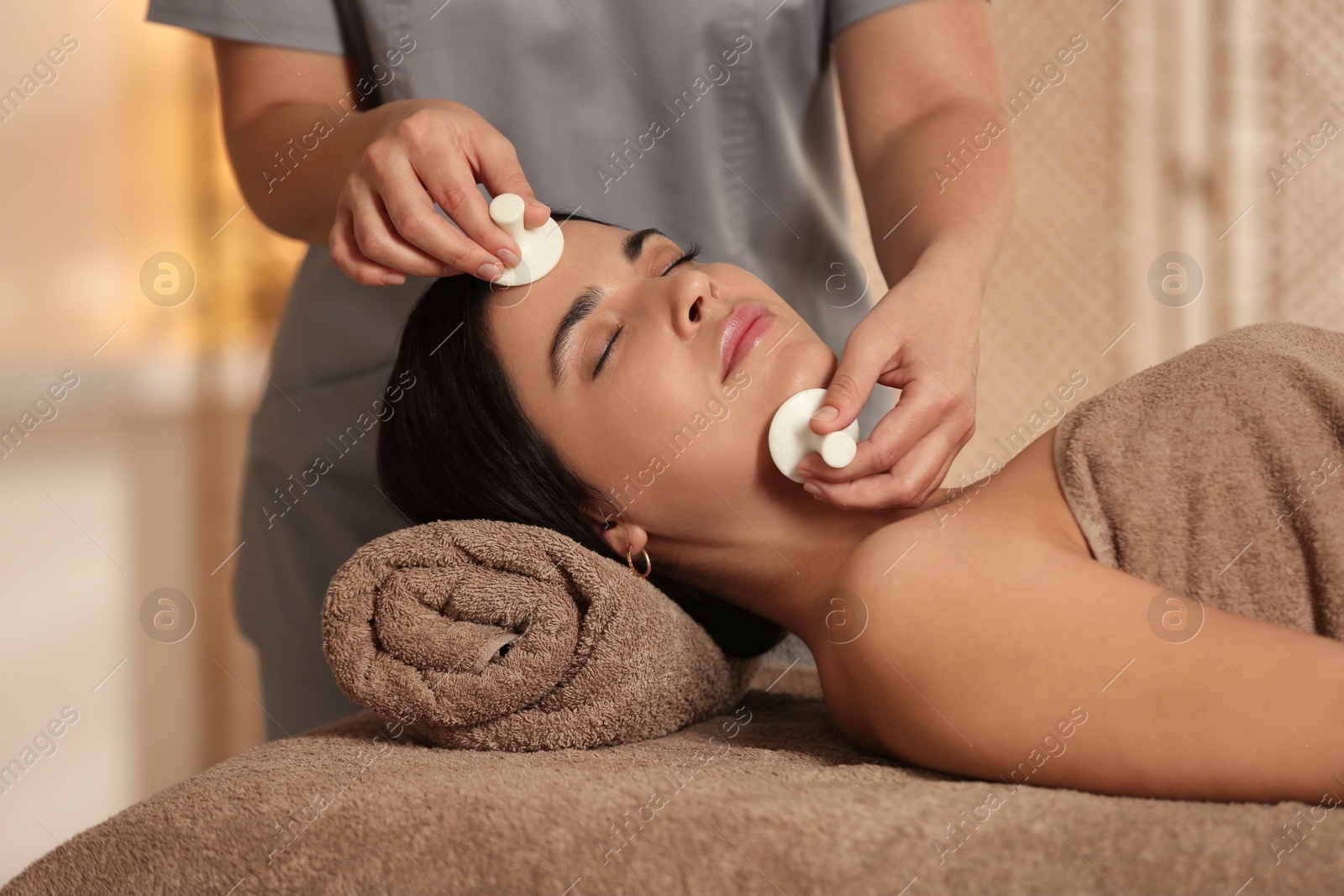 Photo of Young woman receiving facial massage with spa stones in salon