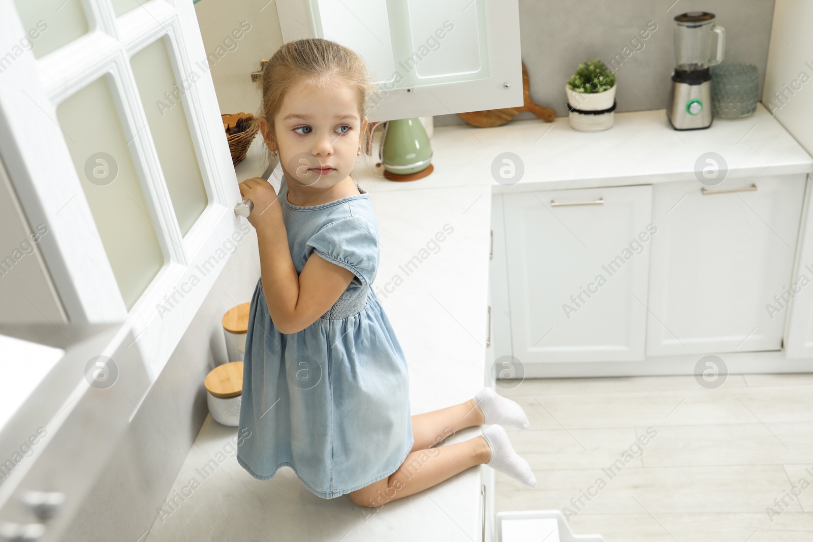 Photo of Little girl sitting on counter and reaching towards shelf in kitchen, above view