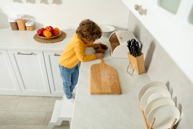 Photo of Little boy with cookies standing on step stool near countertop in kitchen