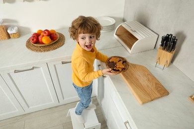 Photo of Little boy with cookies standing on step stool in kitchen