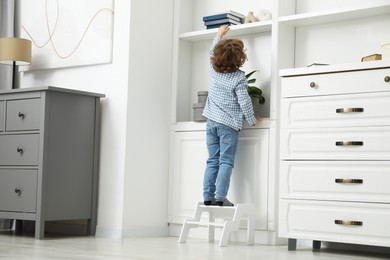 Photo of Little boy standing on step stool and reaching for book at home, back view