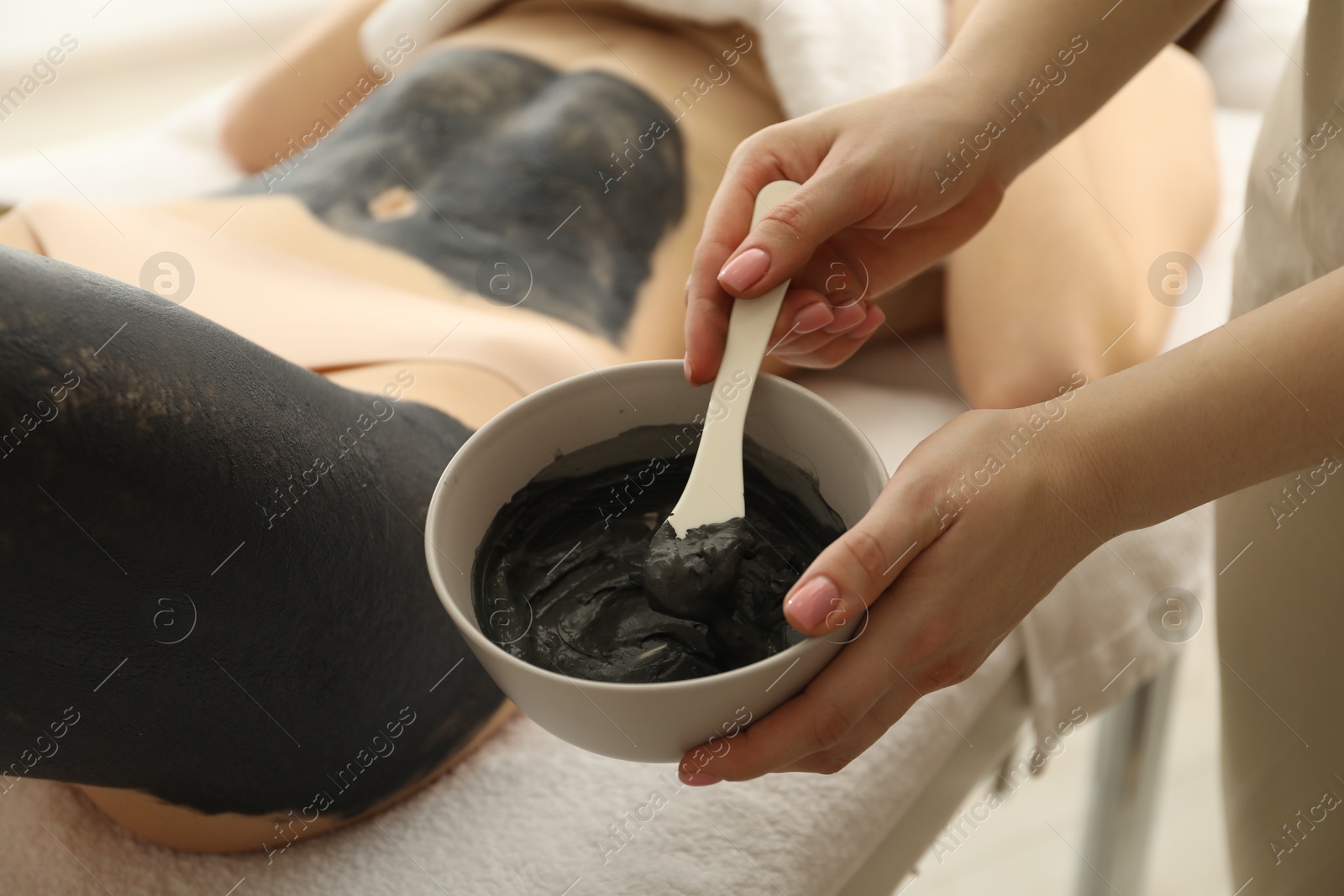 Photo of Esthetician applying cosmetic product for body wraps treatment onto woman's leg in spa salon, closeup