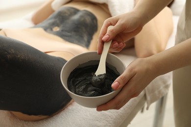 Photo of Esthetician applying cosmetic product for body wraps treatment onto woman's leg in spa salon, closeup