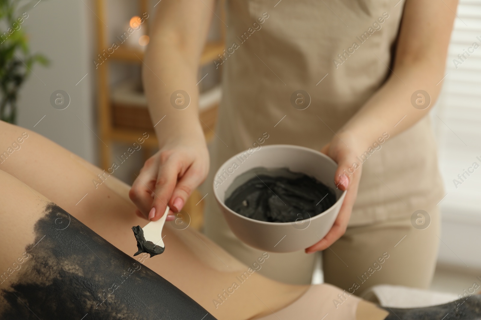 Photo of Esthetician applying cosmetic product for body wraps treatment onto woman's leg in spa salon, closeup