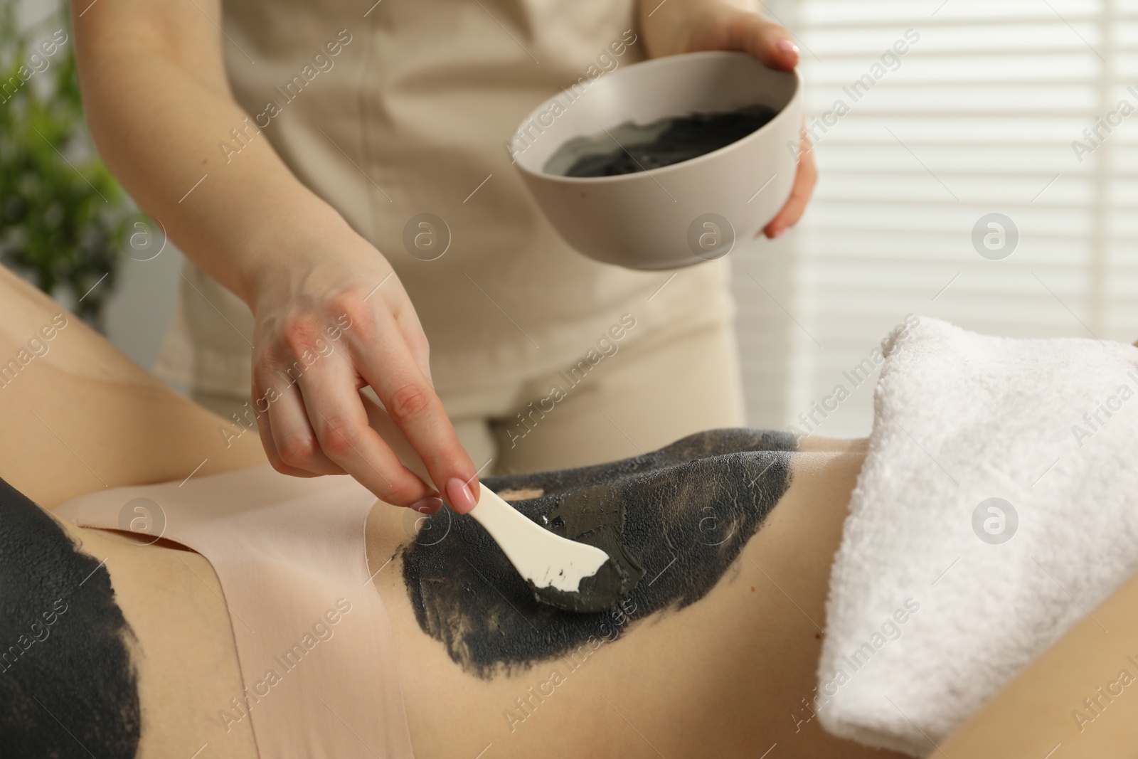 Photo of Esthetician applying cosmetic product for body wraps treatment onto woman's belly in spa salon, closeup