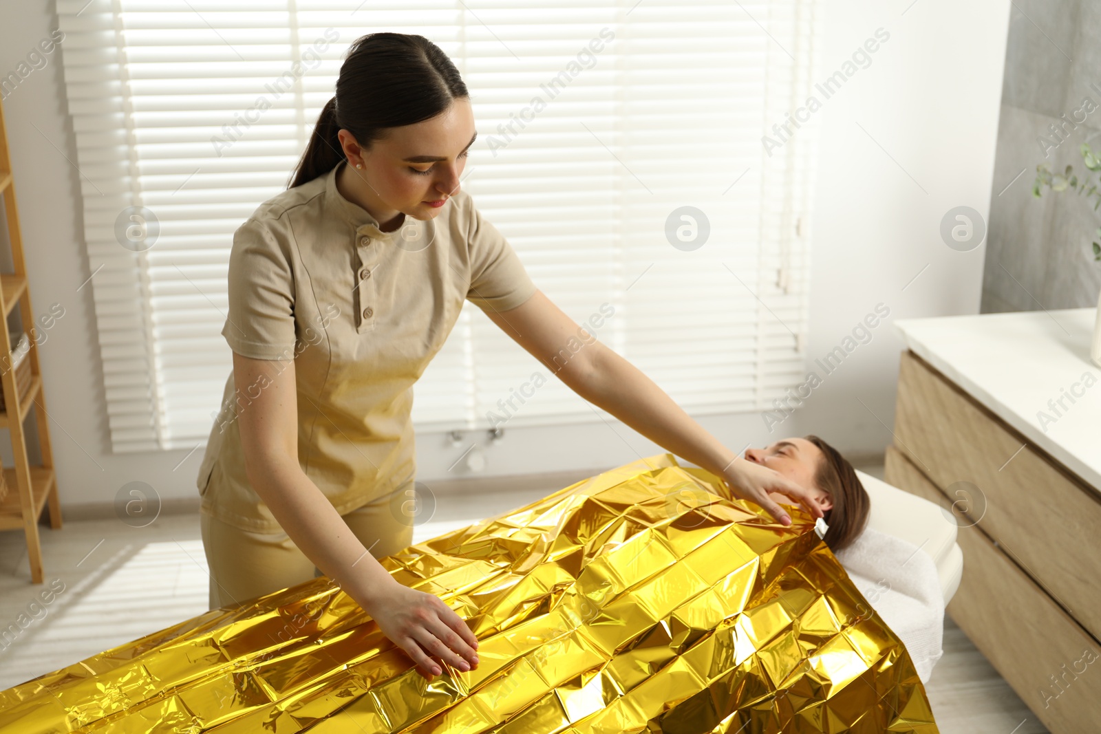 Photo of Spa body wraps. Esthetician covering woman with thermal foil blanket indoors. Space for text