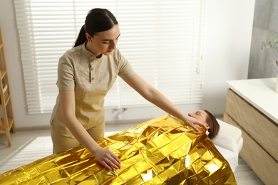 Photo of Spa body wraps. Esthetician covering woman with thermal foil blanket indoors. Space for text