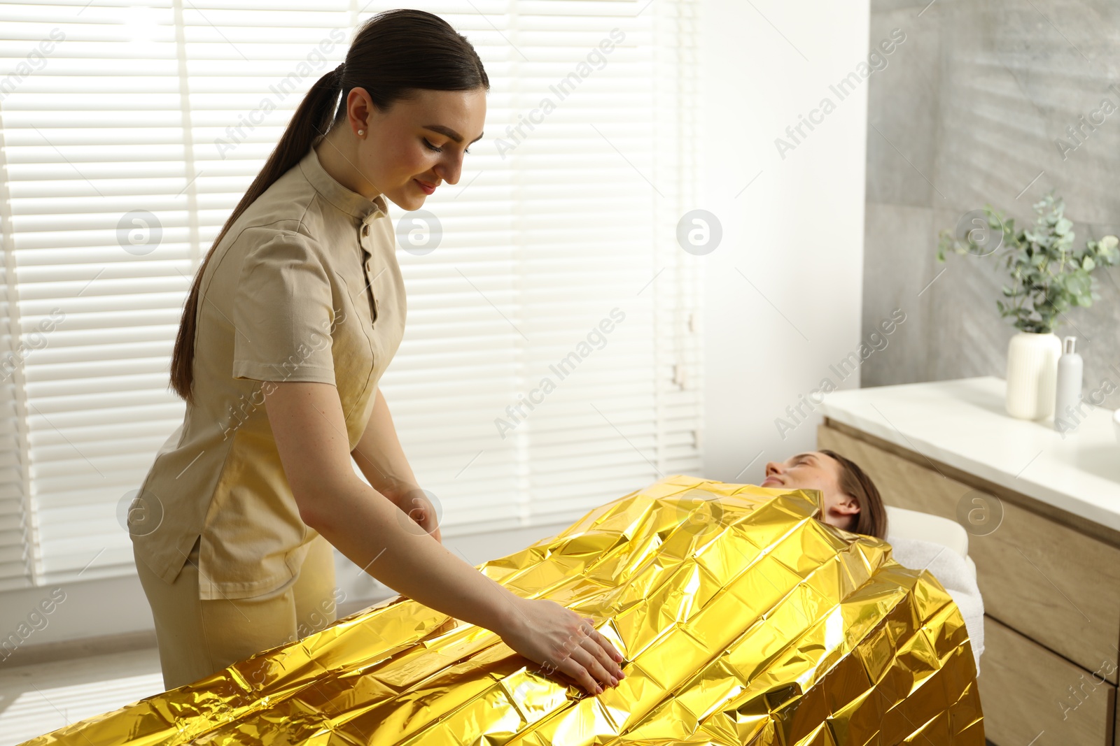 Photo of Spa body wraps. Esthetician covering woman with thermal foil blanket indoors. Space for text