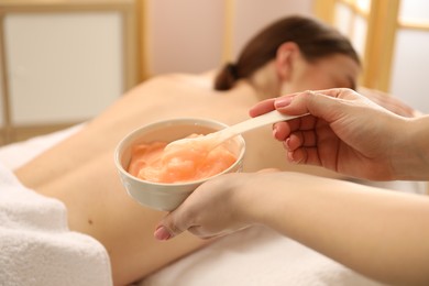 Photo of Esthetician applying cosmetic product for body wraps treatment onto woman's back in spa salon, closeup