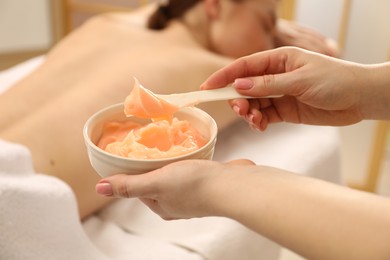 Photo of Esthetician applying cosmetic product for body wraps treatment onto woman's back in spa salon, closeup