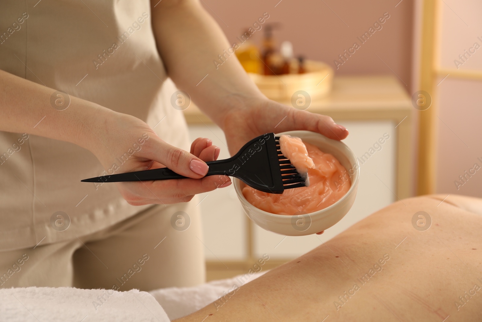 Photo of Esthetician applying cosmetic product for body wraps treatment onto woman's back in spa salon, closeup