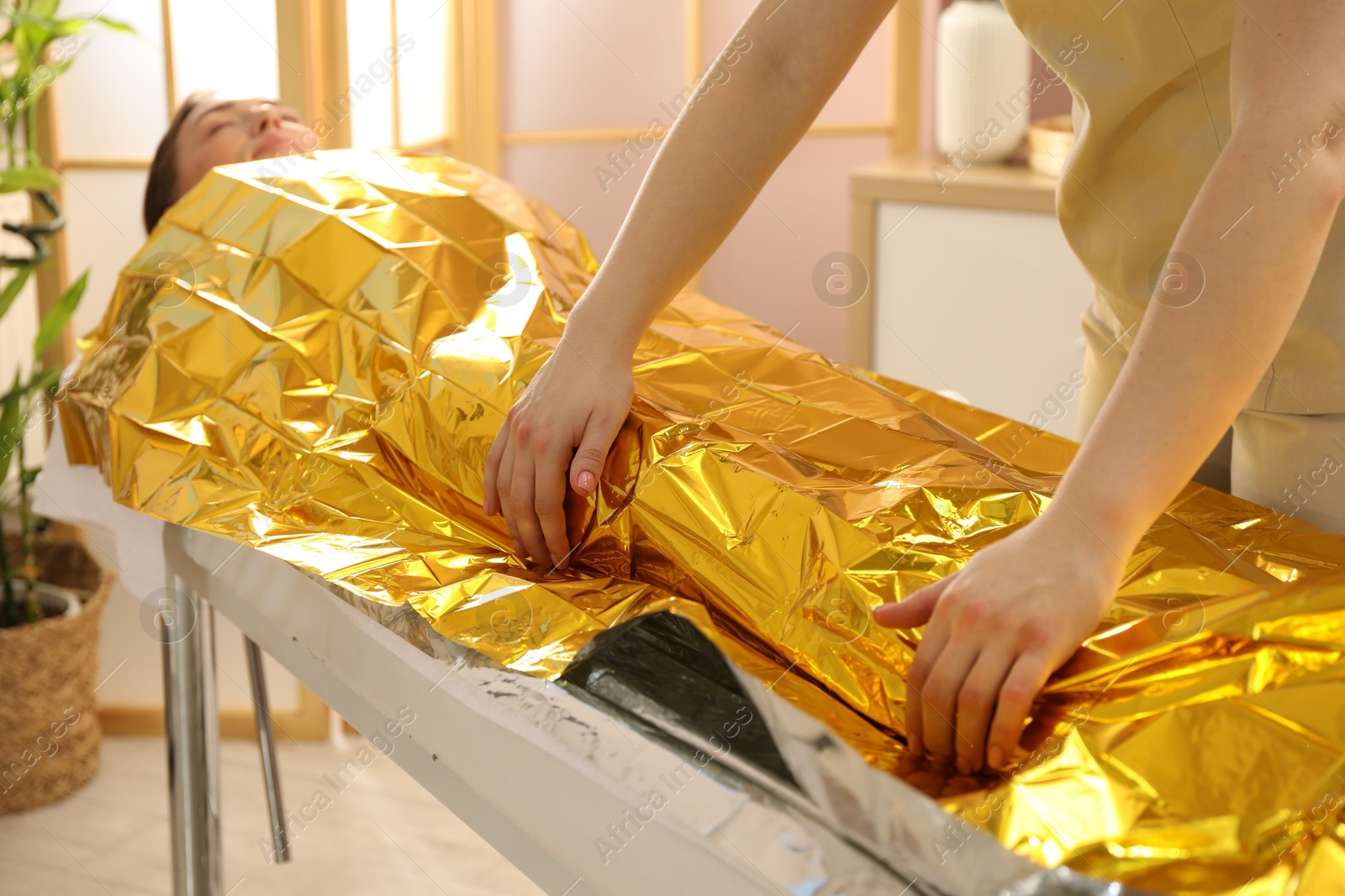 Photo of Spa body wraps. Esthetician covering woman with thermal foil blanket indoors, closeup