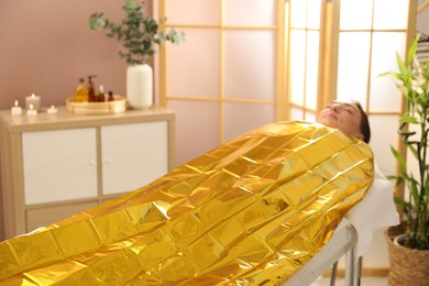 Woman covered with thermal foil blanket undergoing body wraps treatment in spa salon. Space for text