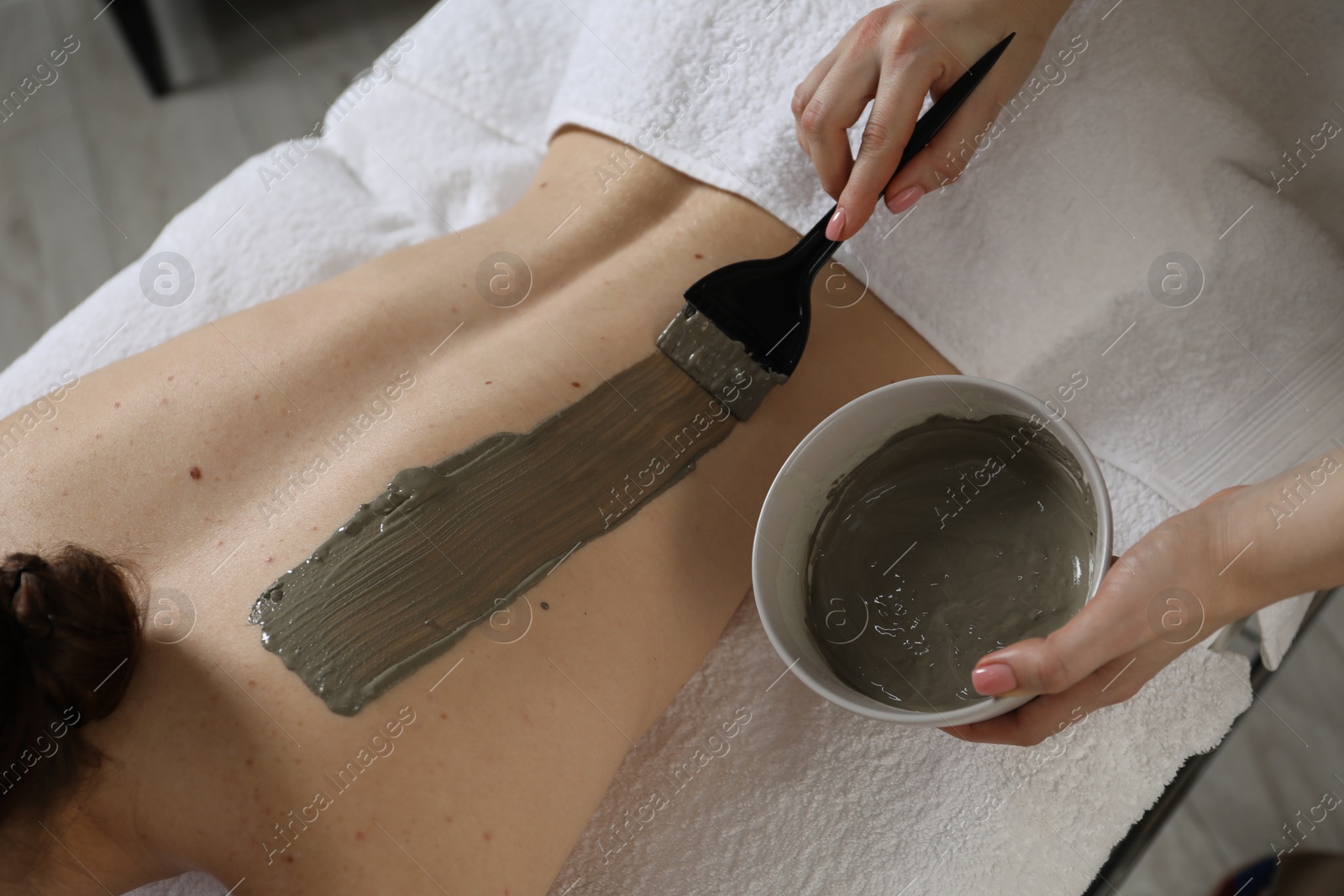 Photo of Esthetician applying cosmetic product for body wraps treatment onto woman's back in spa salon, closeup