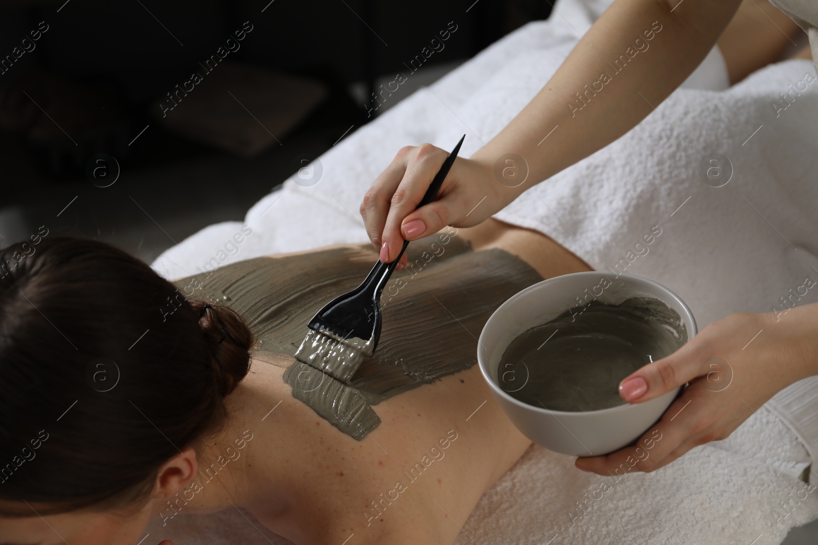 Photo of Esthetician applying cosmetic product for body wraps treatment onto woman's back in spa salon, closeup