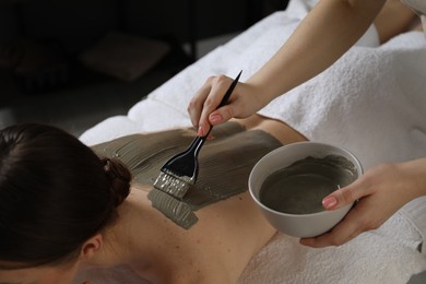 Esthetician applying cosmetic product for body wraps treatment onto woman's back in spa salon, closeup