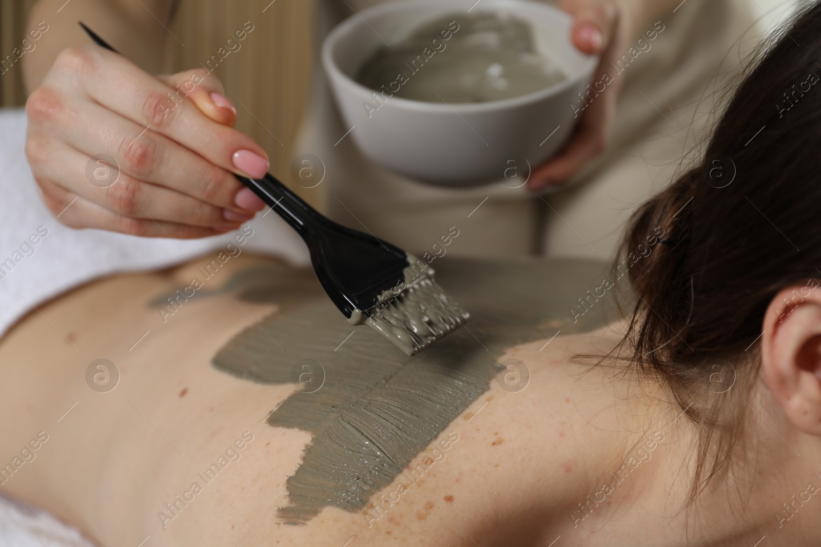 Photo of Esthetician applying cosmetic product for body wraps treatment onto woman's back in spa salon, closeup