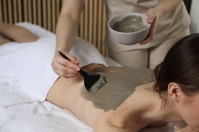 Esthetician applying cosmetic product for body wraps treatment onto woman's back in spa salon, closeup