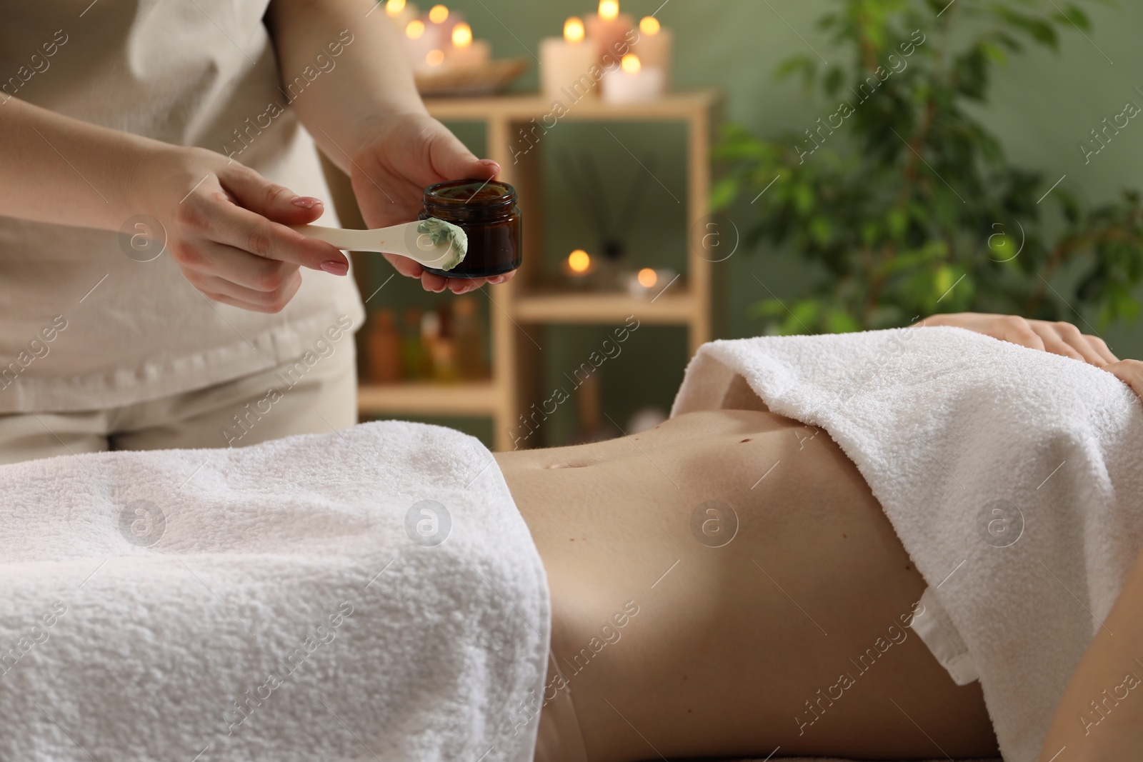 Photo of Esthetician applying cosmetic product for body wraps treatment onto woman's belly in spa salon, closeup