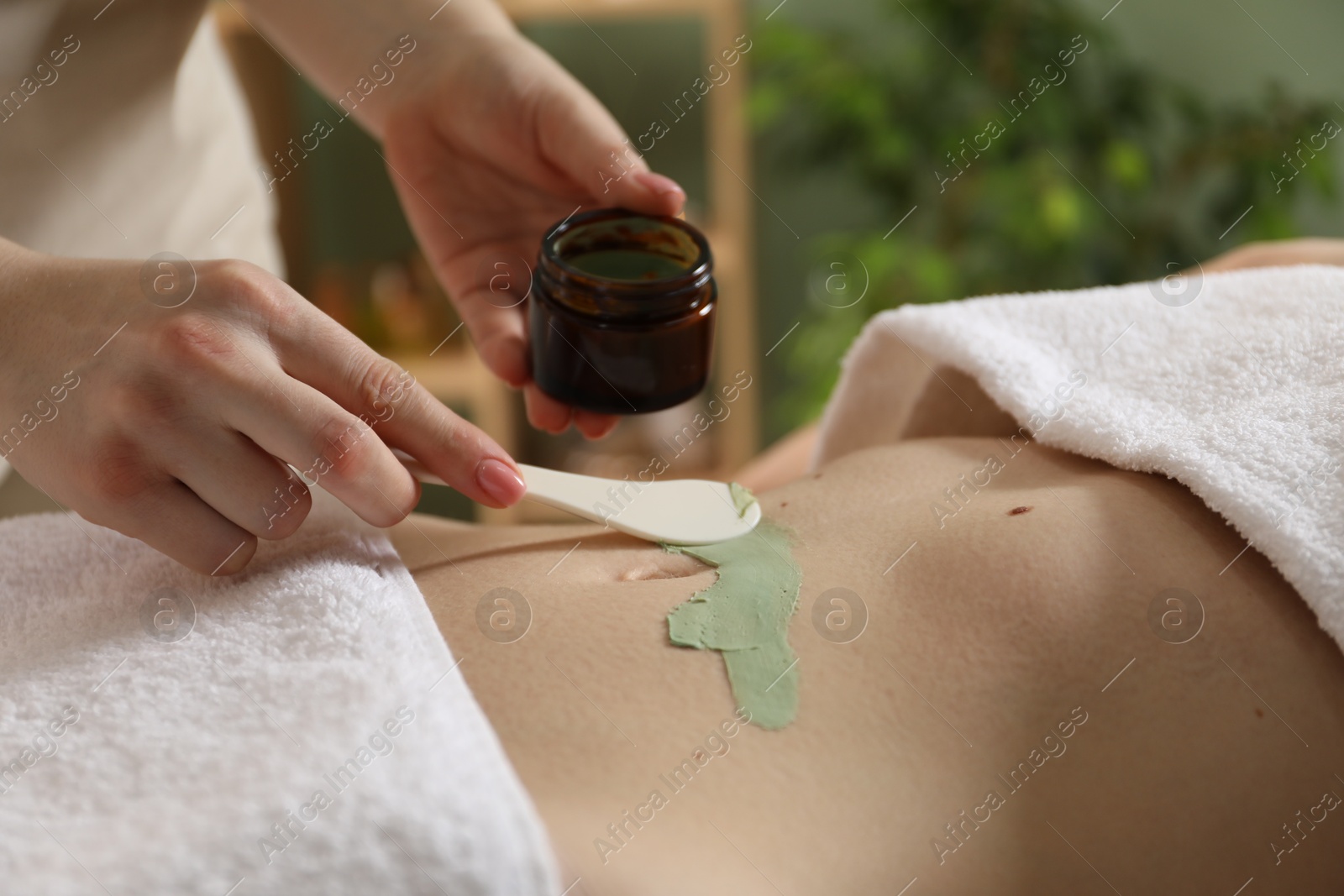 Photo of Esthetician applying cosmetic product for body wraps treatment onto woman's belly in spa salon, closeup