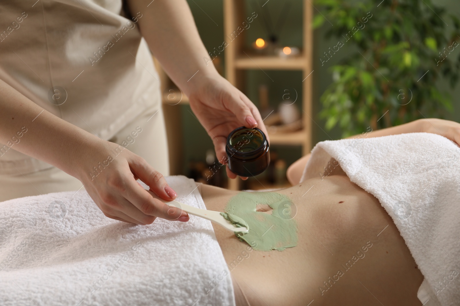 Photo of Esthetician applying cosmetic product for body wraps treatment onto woman's belly in spa salon, closeup