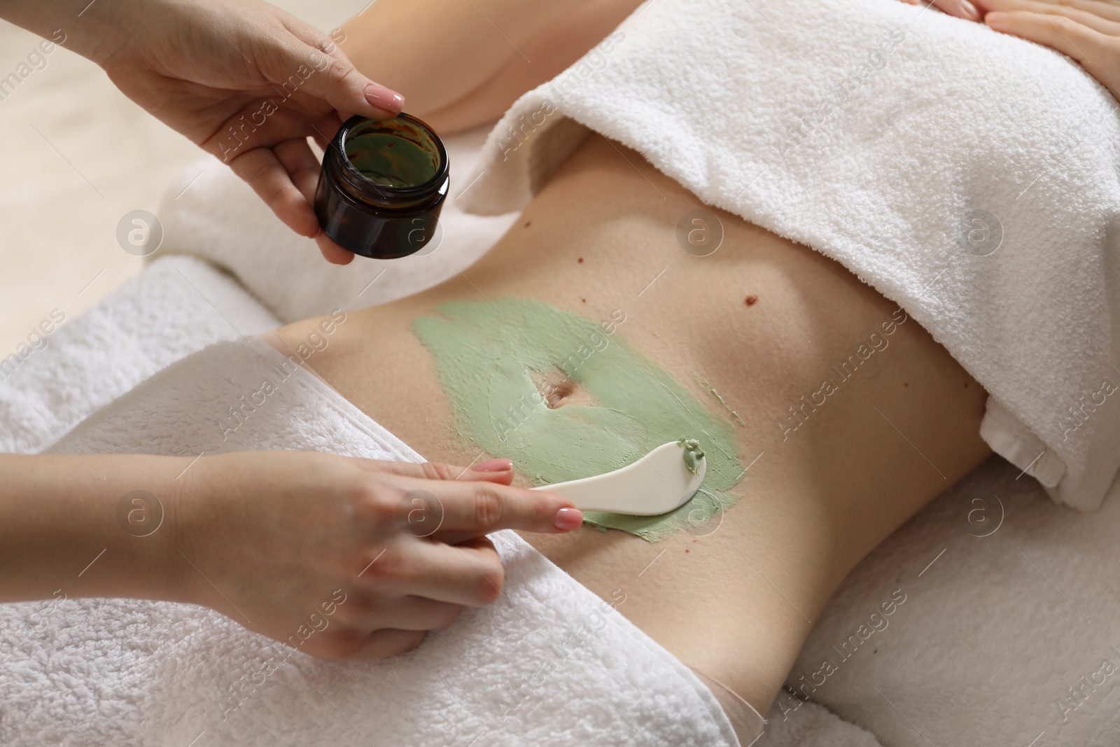 Photo of Esthetician applying cosmetic product for body wraps treatment onto woman's belly in spa salon, closeup