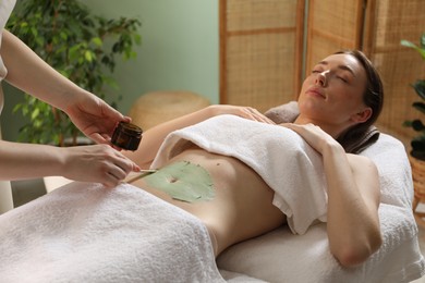 Photo of Esthetician applying cosmetic product for body wraps treatment onto woman's belly in spa salon, closeup