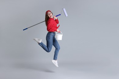 Photo of Woman with roller and bucket of paint jumping on light grey background