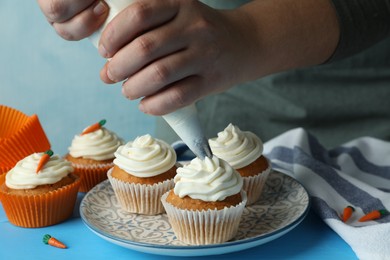 Photo of Woman squeezing whipped cream onto delicious carrot cupcakes at blue table, closeup