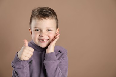 Photo of Cute little boy with missing tooth showing thumbs up on dark beige background, space for text