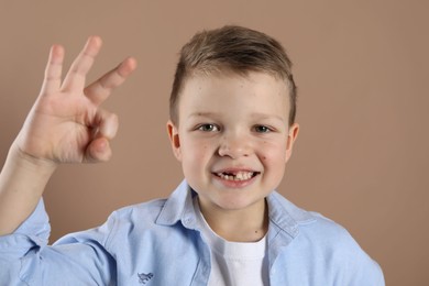 Photo of Cute little boy with missing tooth showing ok gesture on dark beige background