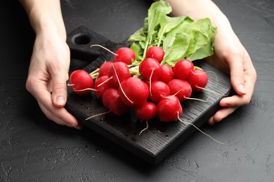 Photo of Woman with board of fresh radishes at dark textured table, closeup