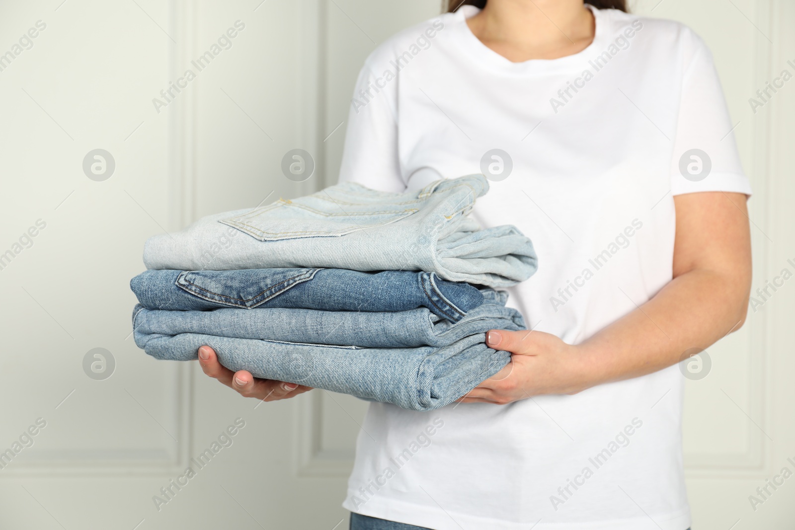 Photo of Woman with stack of different stylish jeans near light wall indoors, closeup