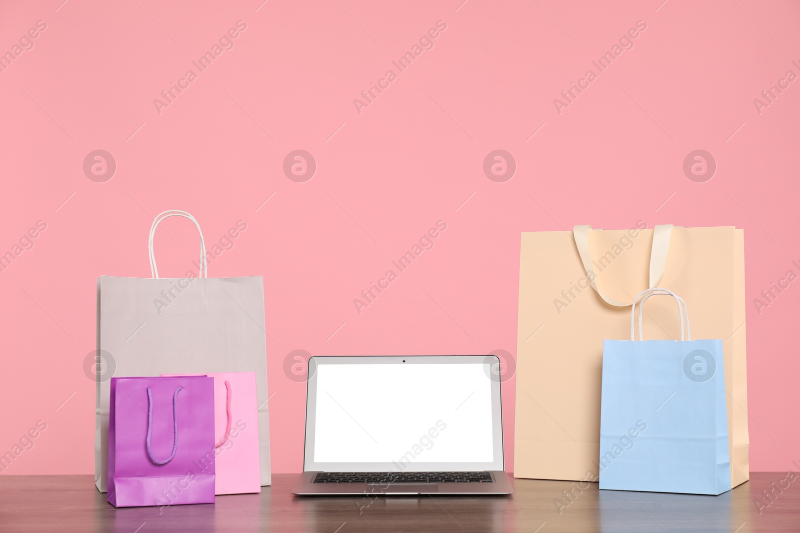 Photo of Internet shopping. Laptop and colorful paper bags on wooden table against pink background