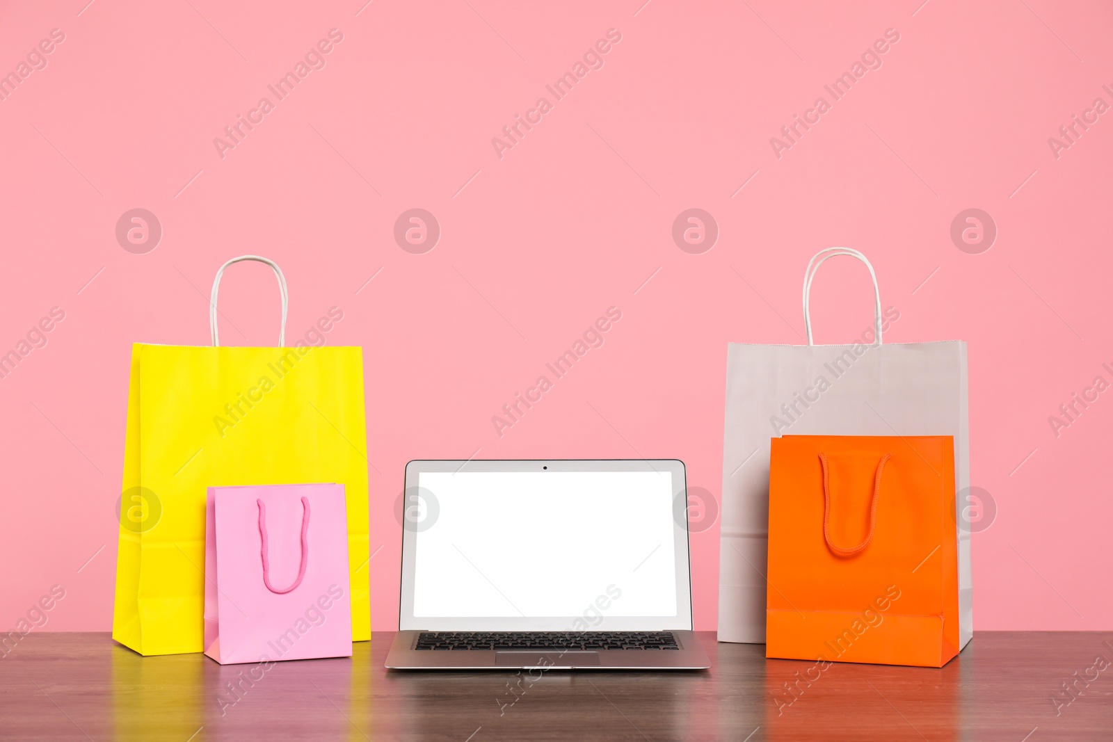 Photo of Internet shopping. Laptop and colorful paper bags on wooden table against pink background