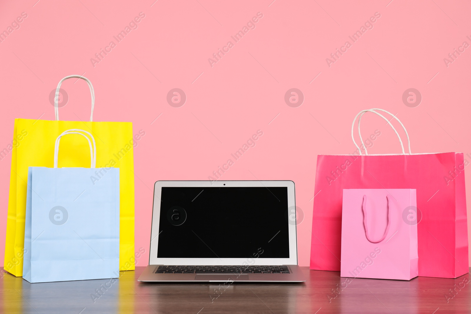 Photo of Internet shopping. Laptop and colorful paper bags on wooden table against pink background