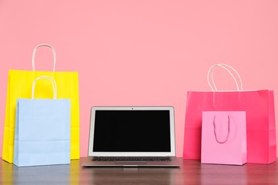 Photo of Internet shopping. Laptop and colorful paper bags on wooden table against pink background