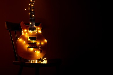 Photo of Electric guitar with Christmas lights on chair against dark background. Space for text