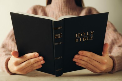 Photo of Woman reading Holy Bible at beige table, closeup