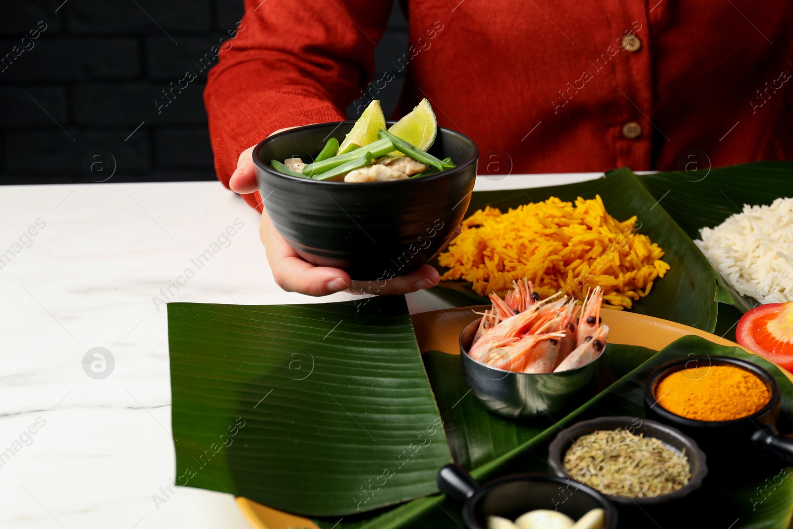 Photo of Woman holding bowl with tasty food at white table with pieces of banana leaves, closeup