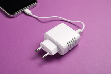 Photo of USB power adapter with charge cable connected to smartphone on purple background, closeup