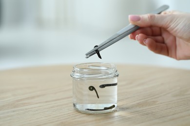 Photo of Woman taking leech from jar with tweezers on wooden table, closeup