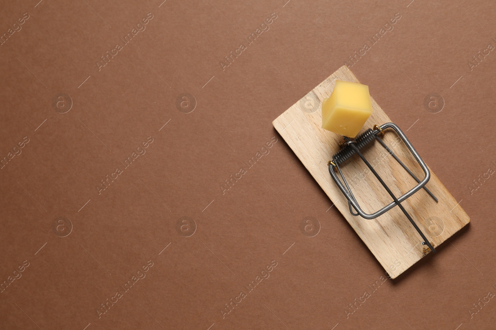 Photo of Spring-loaded bar mousetrap with piece of cheese on brown background, top view. Space for text