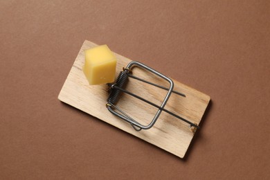 Photo of Spring-loaded bar mousetrap with piece of cheese on brown background, top view